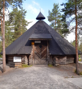 Aarnikotkan kota: suuri, tummakattoinen hirsikota metsäisessä ympäristössä. Rakennuksessa on jyrkkä kartiomainen katto, jonka huipulla on pieni ilmanvaihtorakennelma.