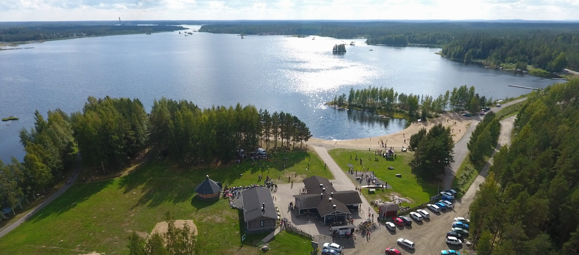 Ilmakuva Kyrkösjärven rannasta, jossa Lakeuden Elämysliikunnan Käpälikkö-rakennus ja hiekkaranta täyttyvät ihmisistä aurinkoisena päivänä. Taustalla avautuu järvimaisema pienine saarineen.