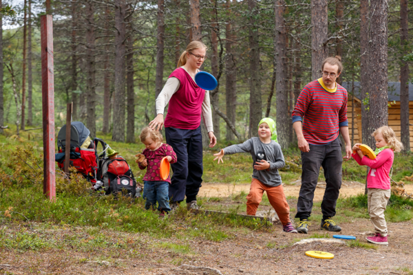 Perhe pelaa frisbeegolfia metsäisessä maastossa. Lapset ja aikuiset heittävät värikkäitä frisbeetä iloisina yhdessä.