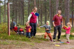 Perhe pelaa frisbeegolfia metsäisessä maastossa. Lapset ja aikuiset heittävät värikkäitä frisbeetä iloisina yhdessä.