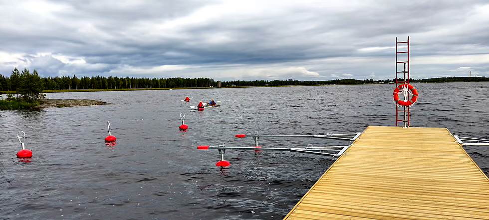 Puiselta laiturilta avautuu näkymä järvelle, jossa on punaisia poijuja ja useita melojia kanootissa. Taustalla näkyy metsäinen rantaviiva ja pilvinen taivas. Laiturin päässä on pelastusrengas ja punainen pelastustikas.